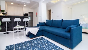 Un salon moderne avec un canapé bleu, un tapis à motifs et un coin cuisine à l’arrière-plan.