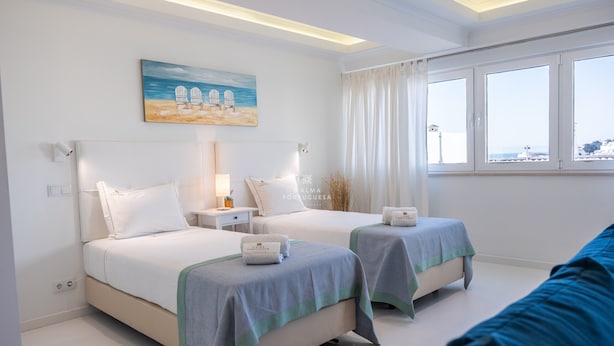 Une chambre d’hôtel avec deux lits, un tableau représentant des chaises de plage, une fenêtre avec des rideaux et une vue sur la ville.