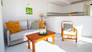 A living room with a sofa, a wooden coffee table, a wicker chair, a lamp, and a kitchen in the background. - Downtown Apartment - By D`alma Portu (Albufeira)