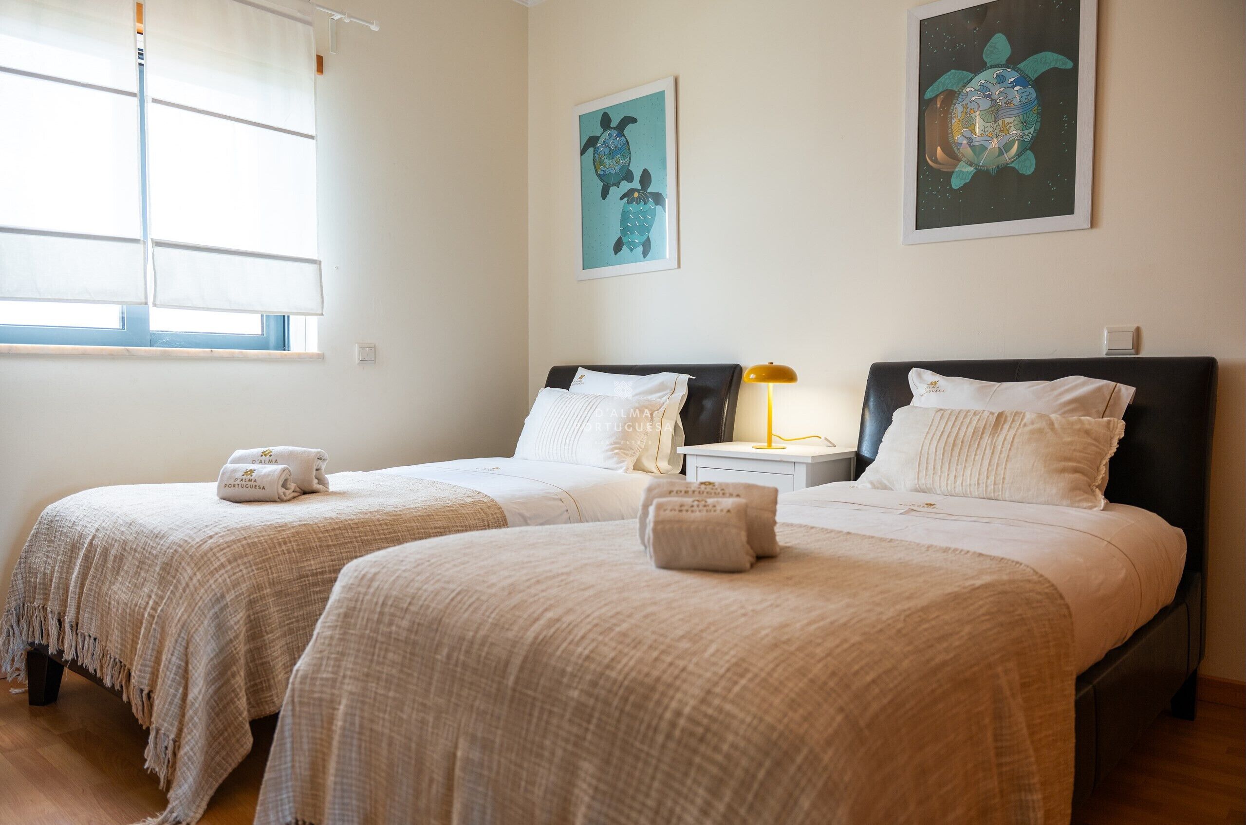 A hotel room with two beds, a window with blinds, a nightstand with a lamp, and artwork on the walls.