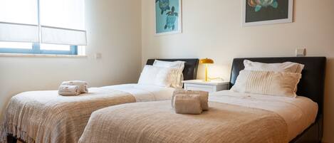 A hotel room with two beds, a window with blinds, a nightstand with a lamp, and artwork on the walls.