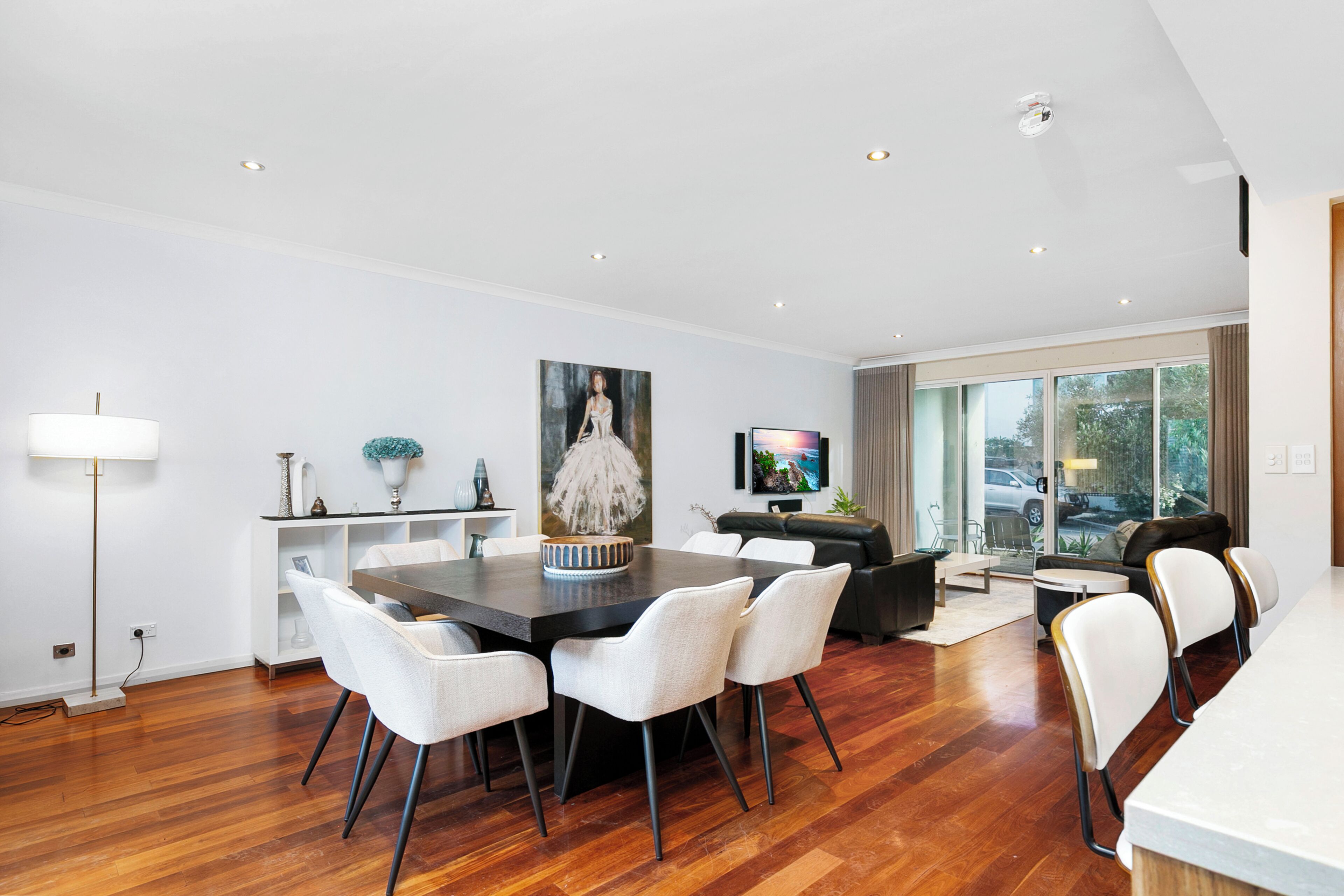 A modern living room with a dining area, featuring a large dining table, white chairs, a sofa, and a TV.