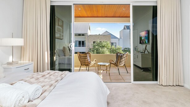 A spacious bedroom with a large bed, a balcony with wicker chairs, and a view of the outside. - Walk to Beach Coastal Home in Coogee (Perth)