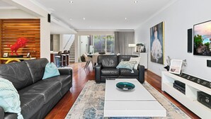 A modern living room with a black leather sofa, a white coffee table, and a dining area in the background. - Walk to Beach Coastal Home in Coogee (Perth)