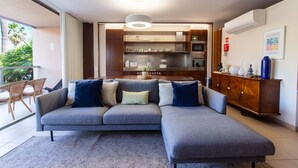 A modern living room with a grey sectional sofa, a wooden cabinet, and a kitchen area with stainless steel appliances. - Apartamento Dunas Mar (Albufeira)