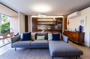 Ein modernes Wohnzimmer mit einem grauen Ecksofa, einem Holzschrank und einer Küchenzeile mit Edelstahlgeräten.