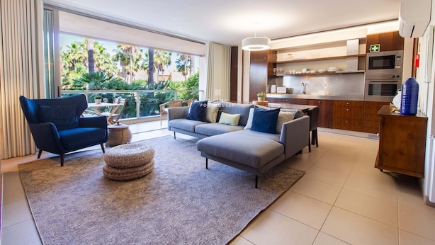 A modern living room with a sofa, armchair, and ottoman, a kitchen with appliances, and a balcony with plants. - Apartamento Dunas Mar (Albufeira)