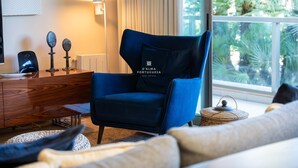 A modern living room with a blue armchair, a wooden dresser, and a view of greenery outside. - Apartamento Dunas Mar (Albufeira)
