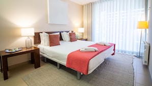 A hotel room with a bed, bedside tables, a lamp, a large window with curtains, and a wall-mounted artwork. - Apartamento Dunas Mar (Albufeira)