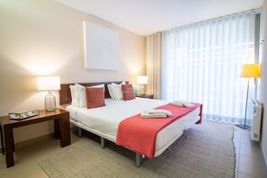 Ein Hotelzimmer mit Bett, Nachttischen, einer Lampe, einem großen Fenster mit Vorhängen und einem Wandbild.