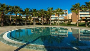A circular pool with a tiled edge, surrounded by palm trees and a building with multiple floors and balconies. - Apartamento Dunas Mar (Albufeira)
