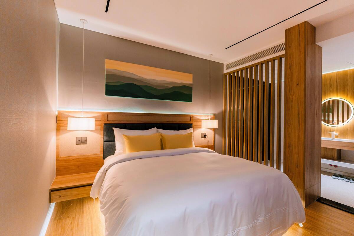 A modern hotel room with a large bed, wooden headboard, bedside lamps, and a wall-mounted artwork.