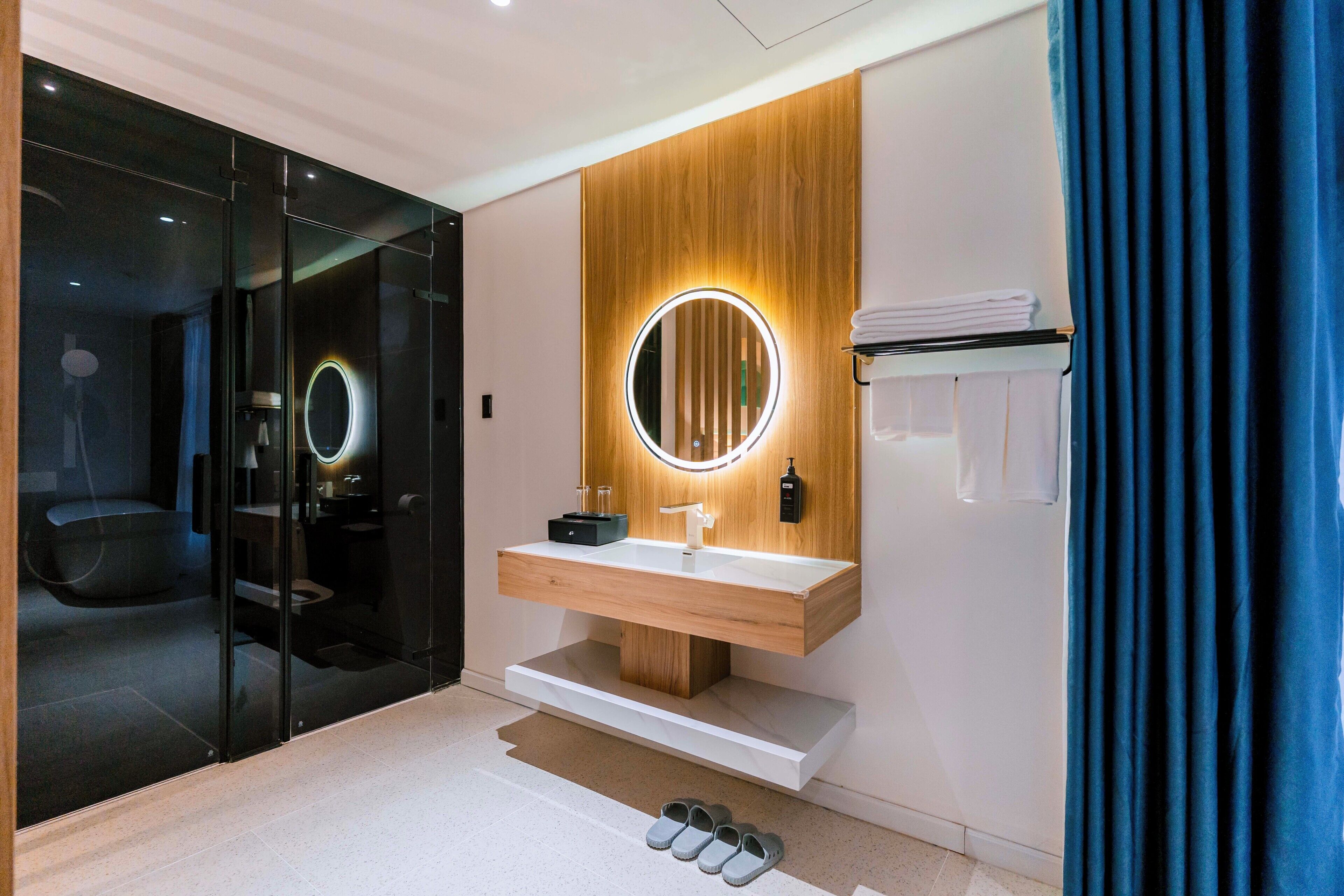 A modern bathroom with a wooden vanity, a round mirror with a light, and a glass shower enclosure.