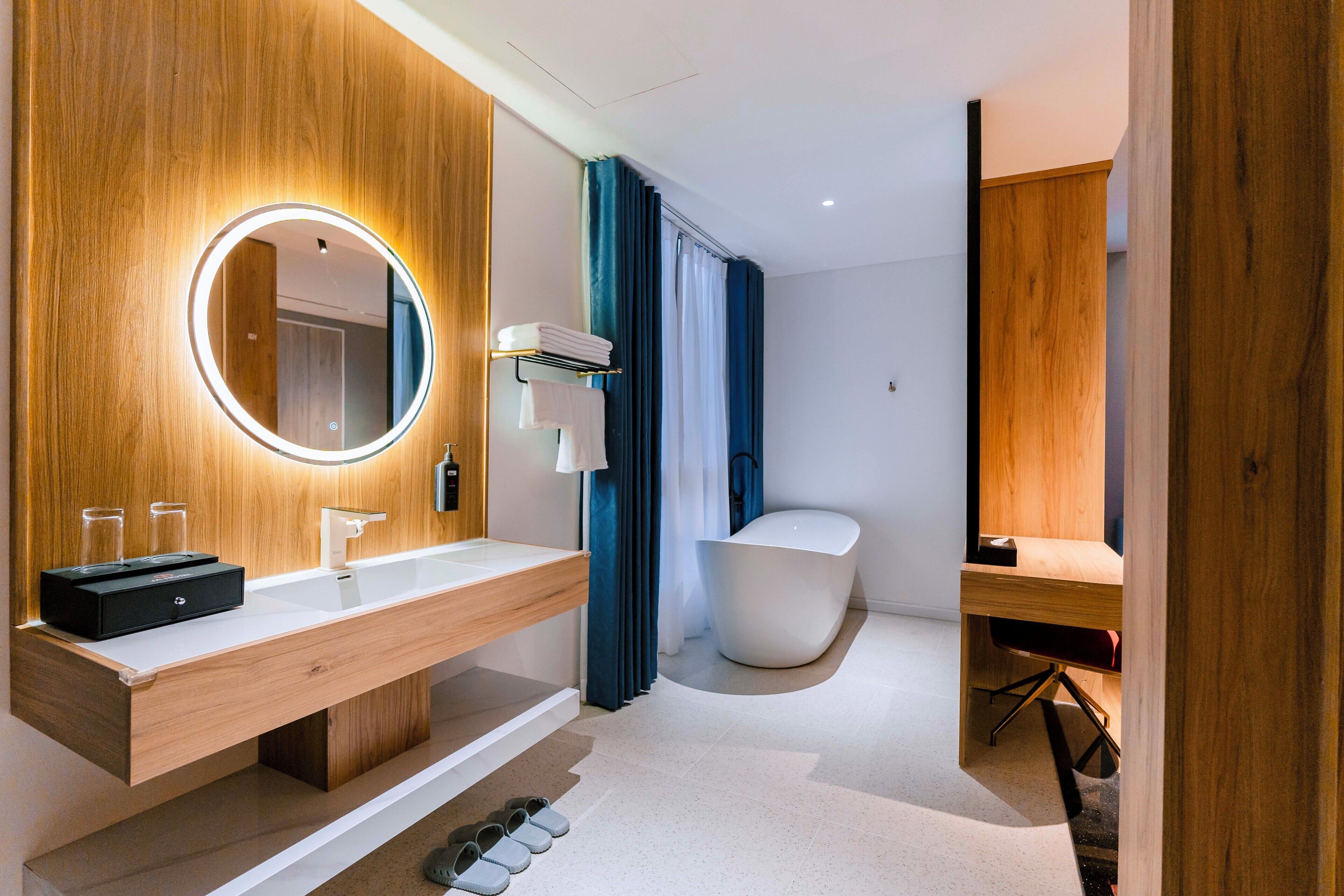 A modern bathroom with a wooden vanity, a freestanding bathtub, and a round mirror with a light.