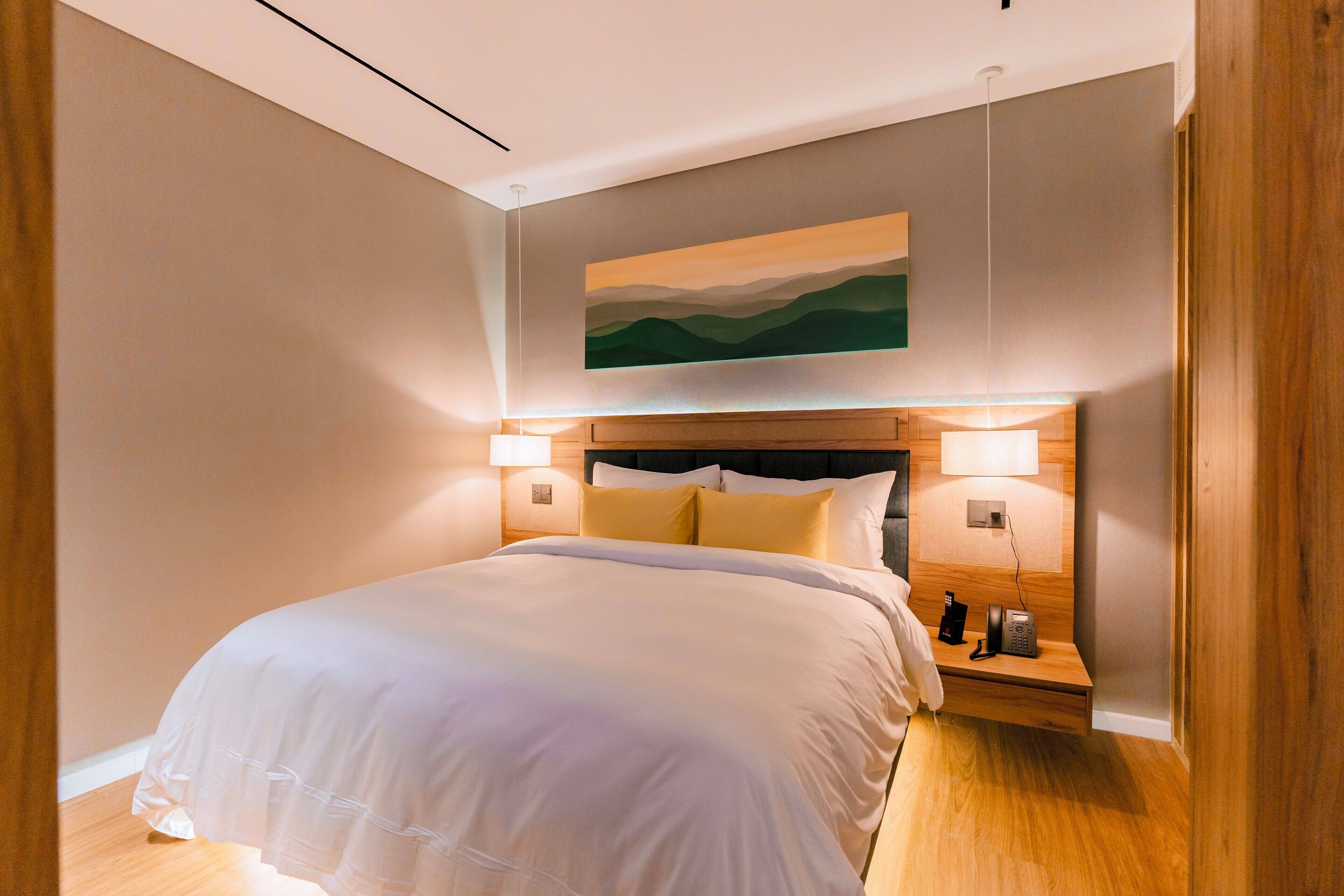 A modern hotel room with a large bed, wooden headboard, bedside lamps, and a painting on the wall.