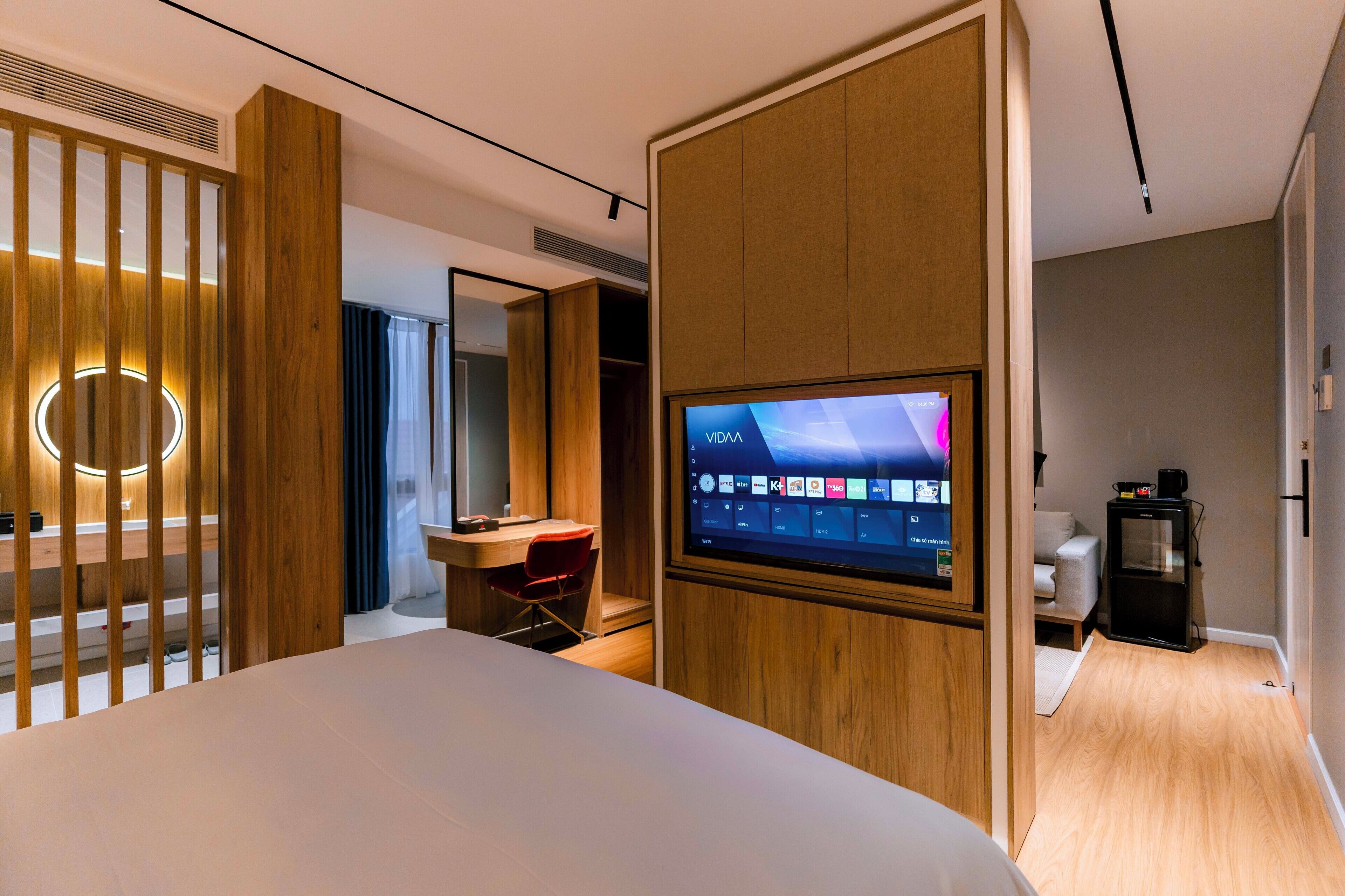 Ein modernes Hotelzimmer mit einem großen Bett, einem an einer Holzvertäfelung befestigten Fernseher, einem Schreibtisch mit Stuhl und einer kleinen Küchenzeile.