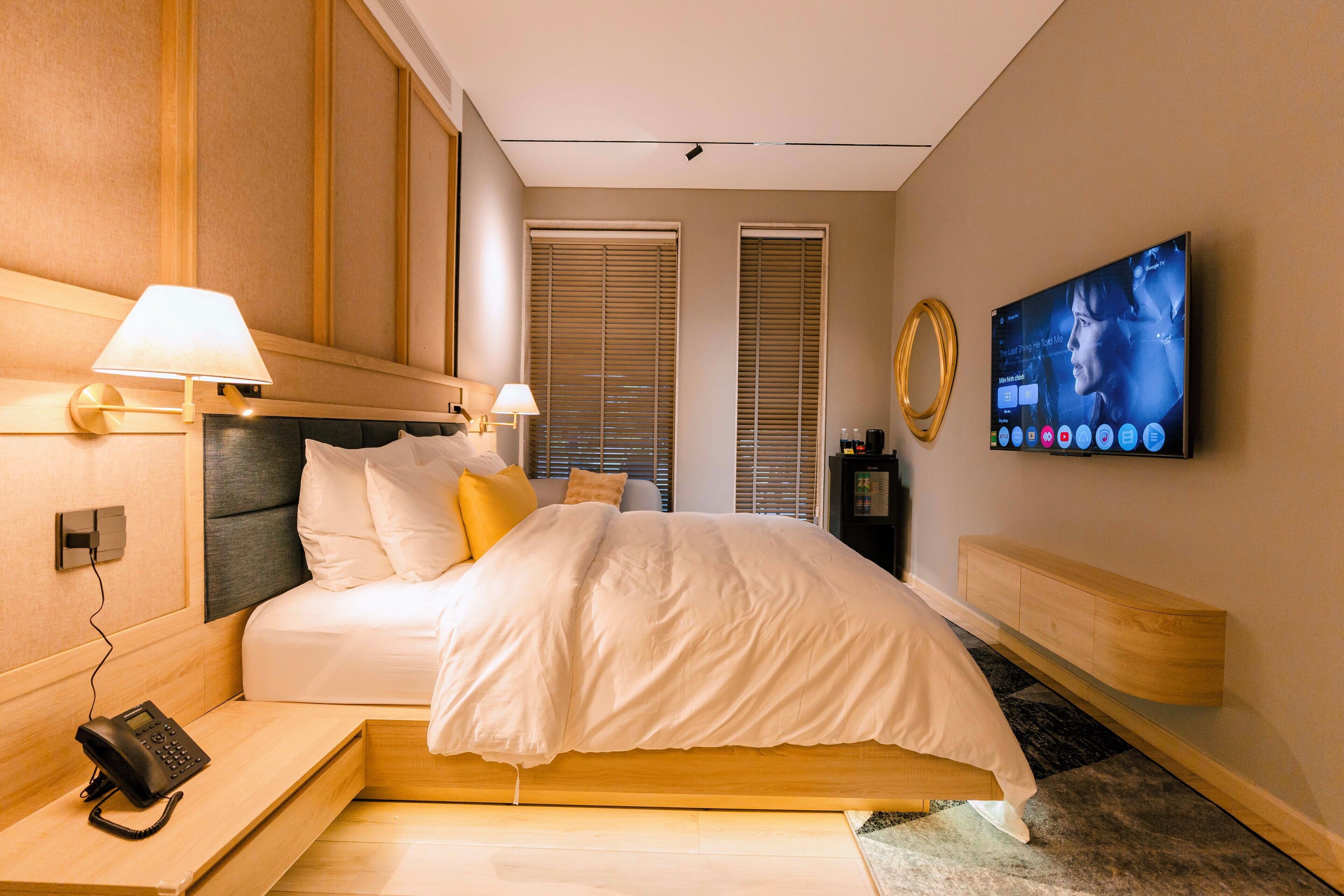 A modern hotel room with a bed, bedside lamps, a mounted TV, a telephone, and a wooden nightstand.