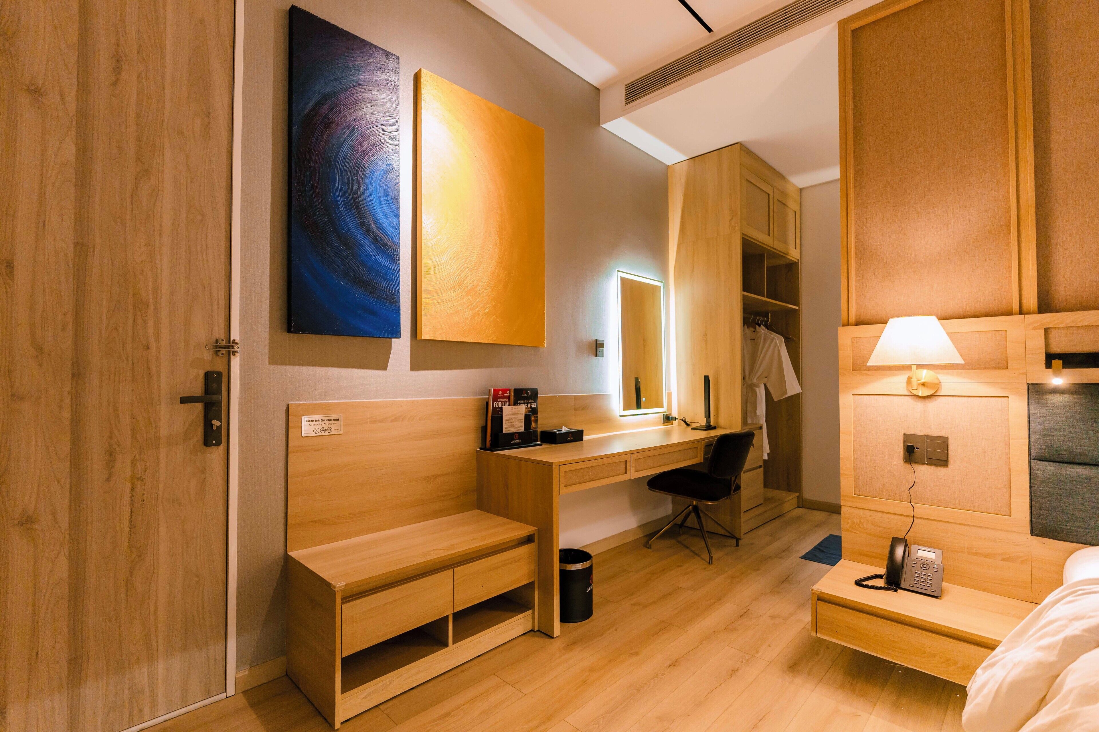A modern hotel room with a wooden desk, a bench, a bed, and a wardrobe.