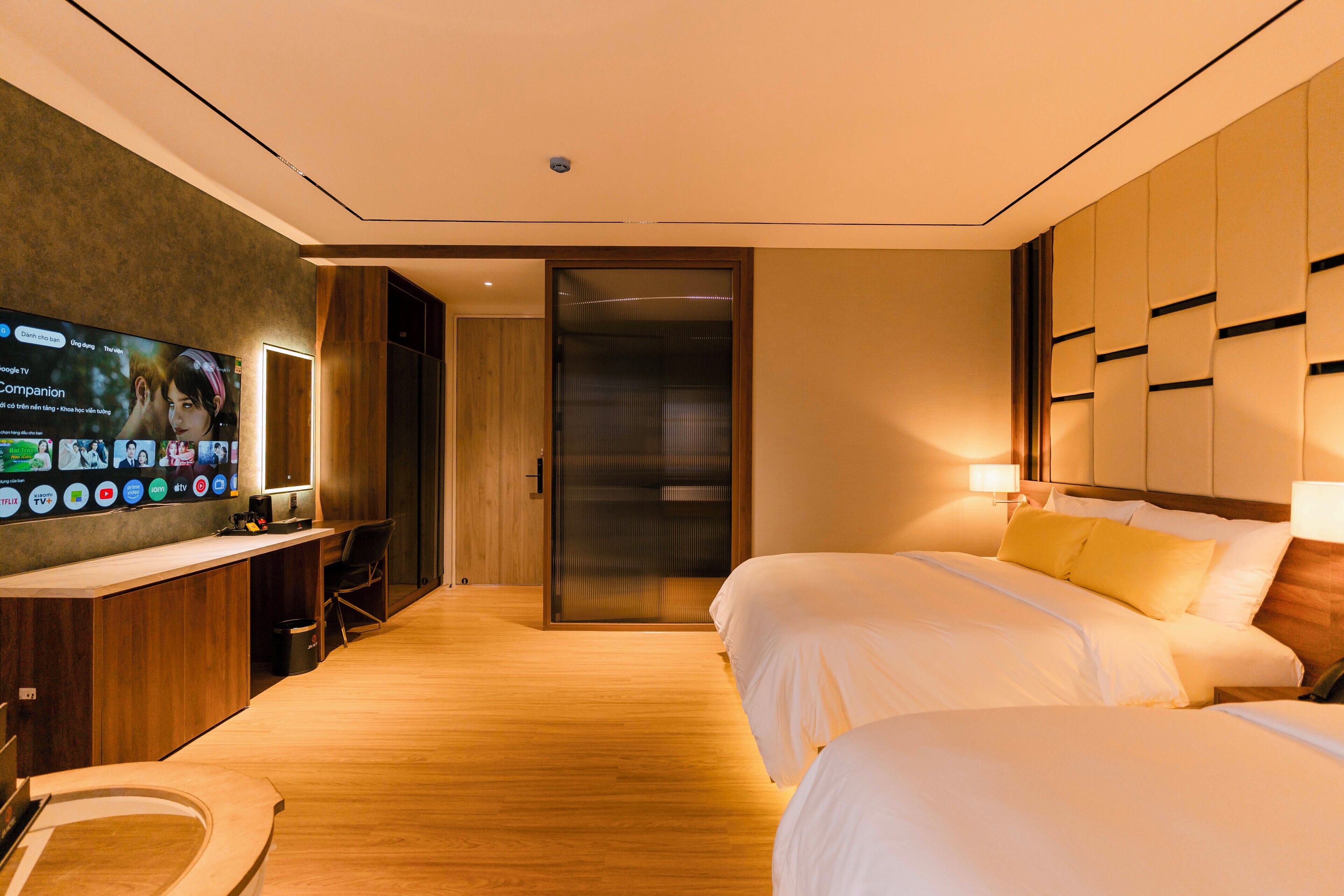 Een hotelkamer met een houten vloer, een aan de muur bevestigde televisie, een bureau met stoel en twee bedden met witte lakens.