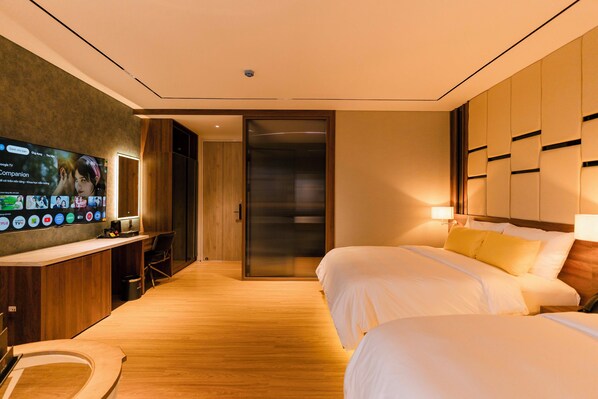 Habitación de hotel con piso de madera, un televisor en la pared, un escritorio con silla y dos camas con ropa blanca.