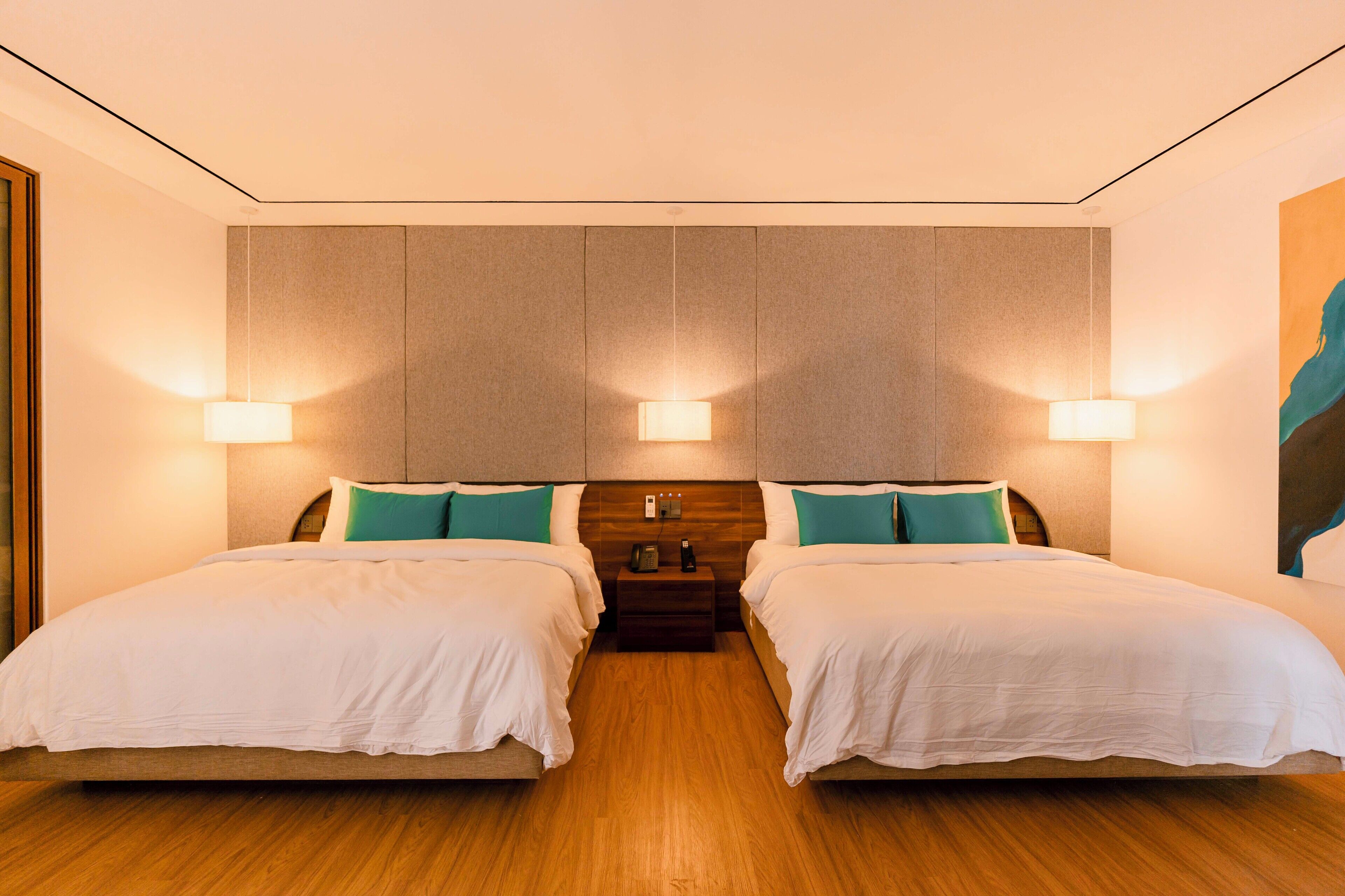 A hotel room with two beds, wooden flooring, a neutral color scheme, and wall-mounted lighting.