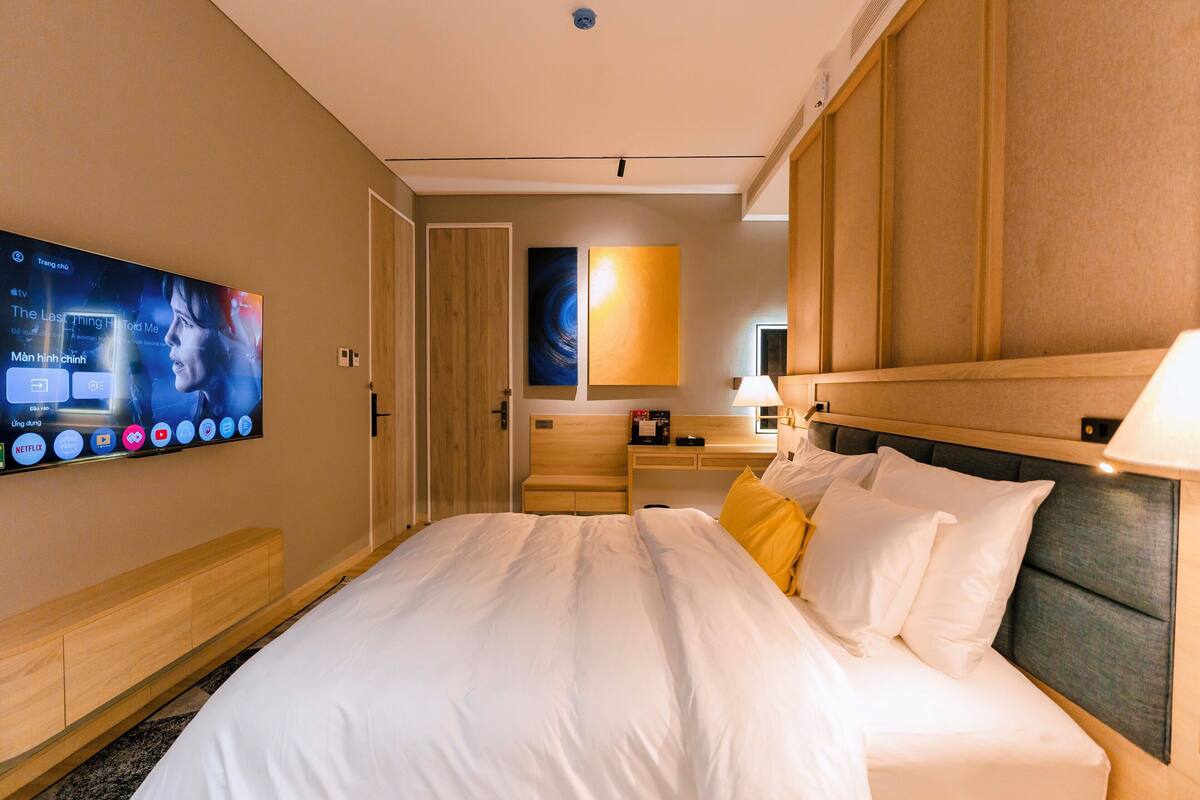A hotel room with a large bed, a TV mounted on the wall, a wooden headboard, and a bedside table.