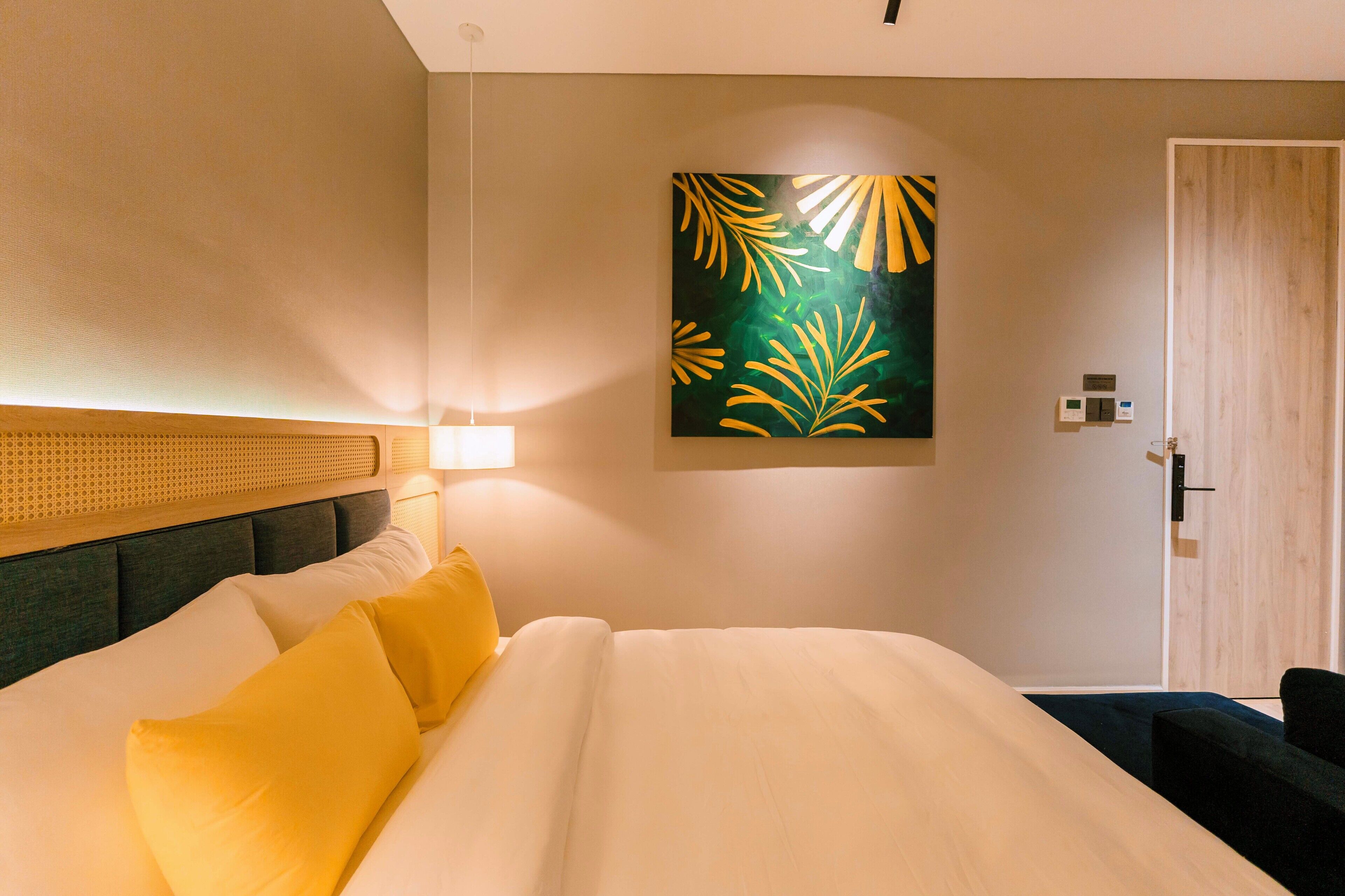 A hotel room with a bed, a headboard, a wall-mounted artwork, a door, and a nightstand with a lamp.