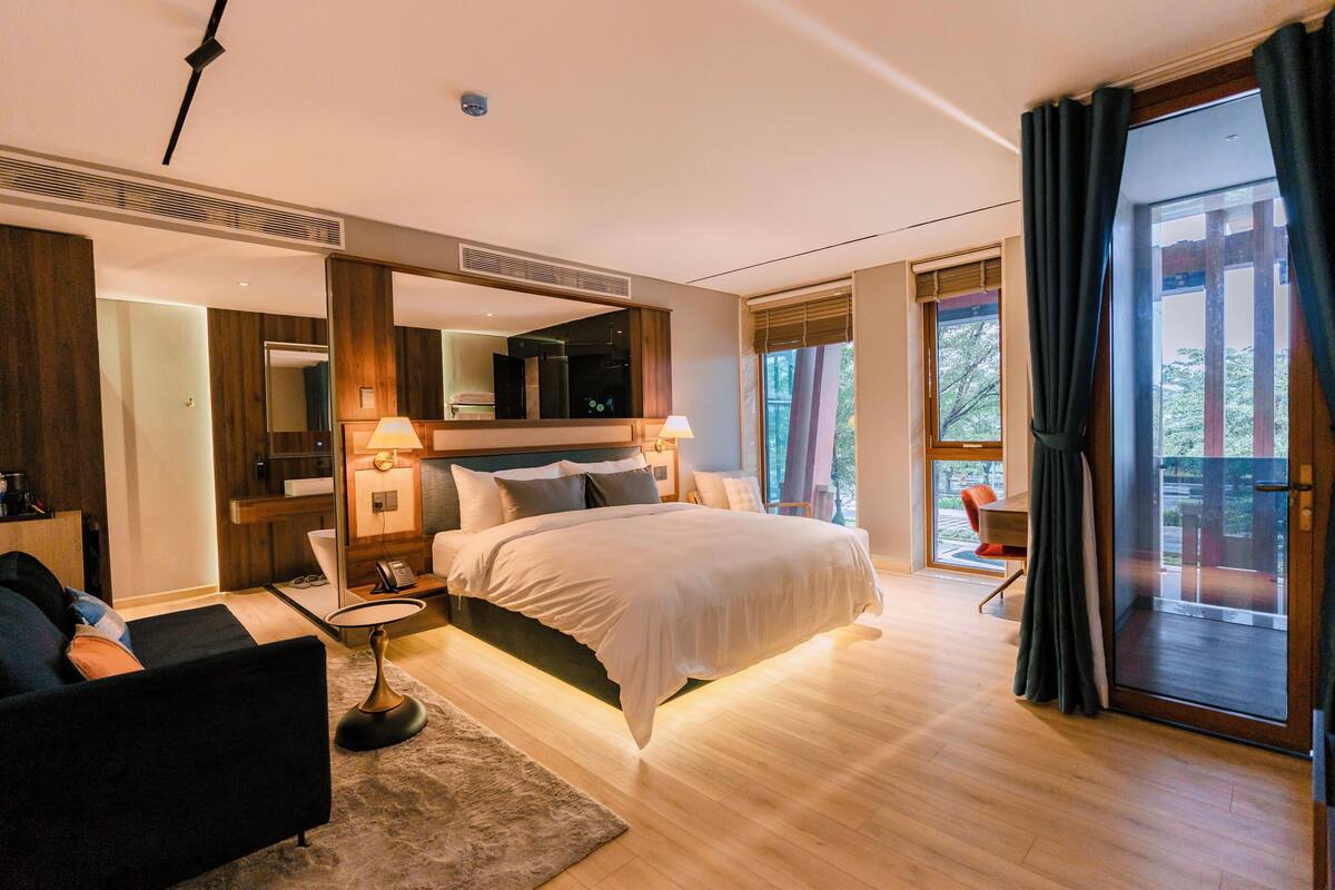 A modern bedroom with a large bed, a bedside table, a chair, and a small table. There is a large window with curtains and a view of trees.