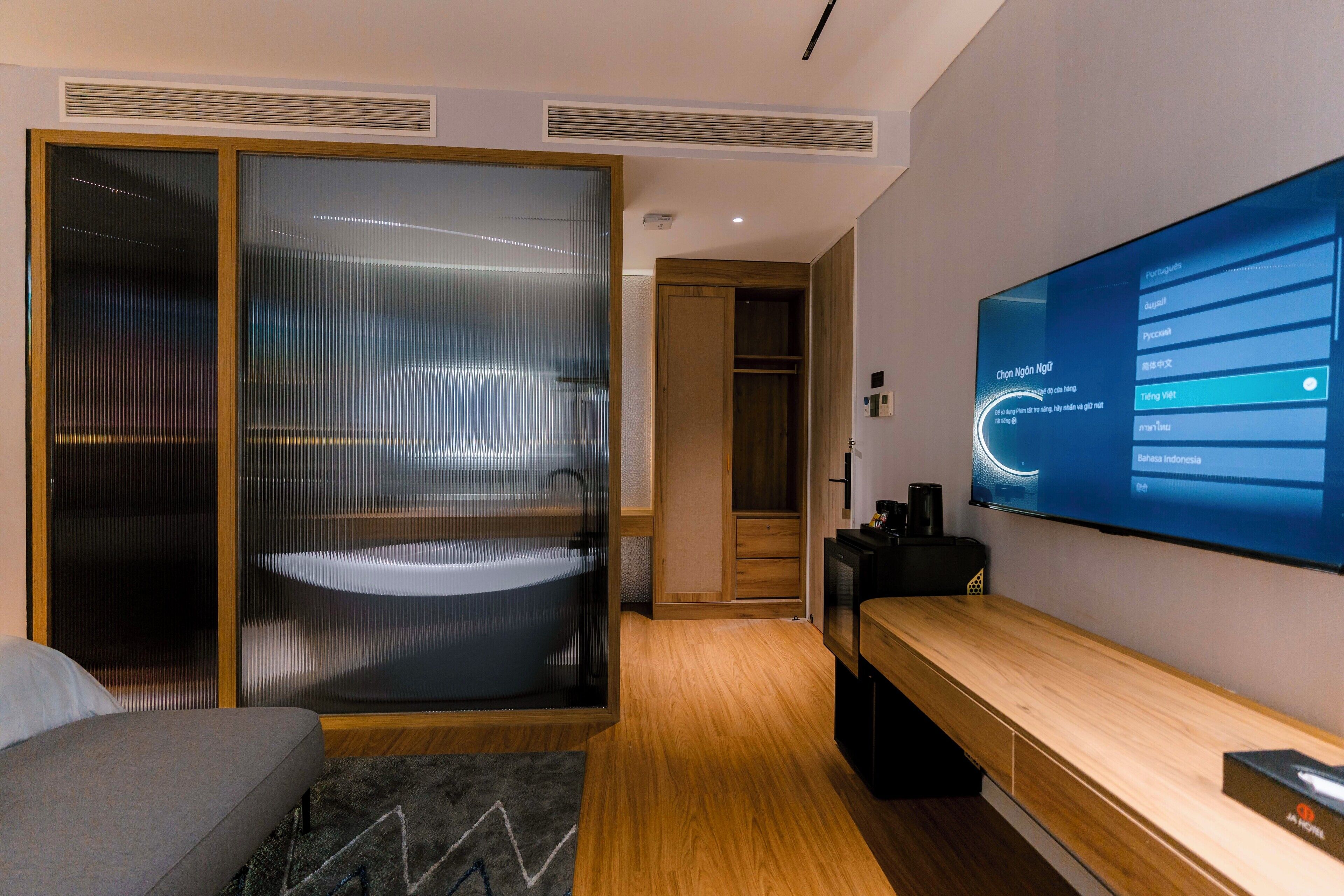 A modern hotel room with a large TV, a sliding glass door closet, and a wooden bench.