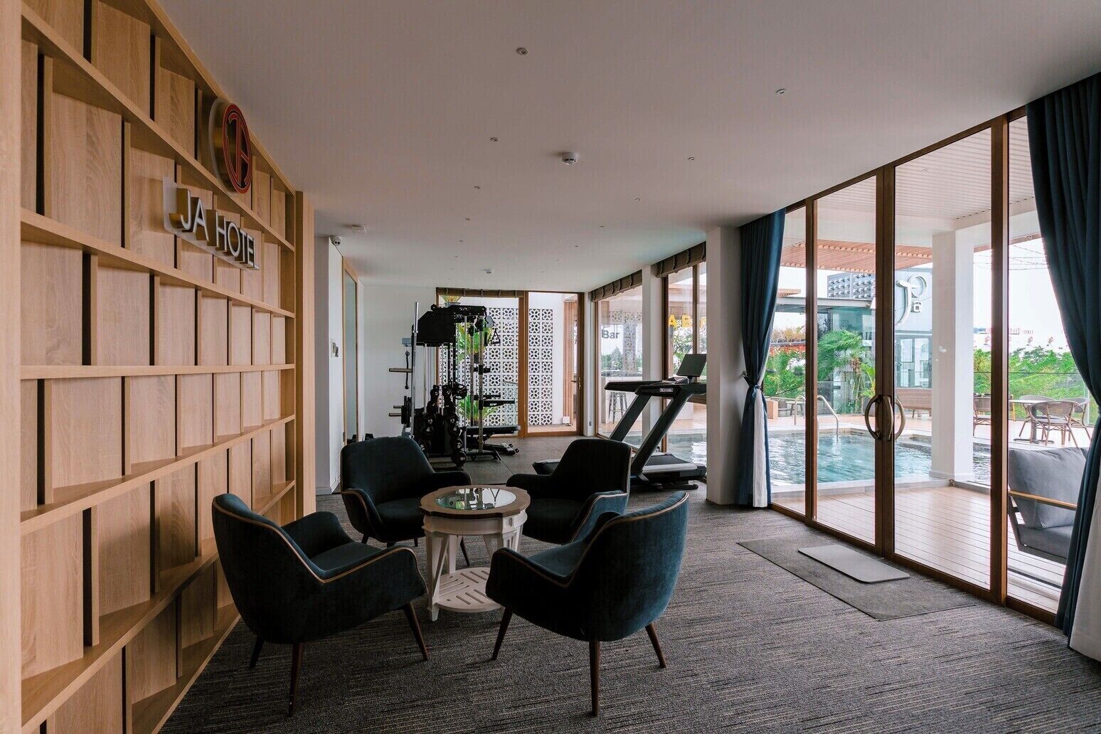 A modern hotel lobby with a wooden wall, a treadmill, exercise equipment, and a pool area visible through large glass doors.