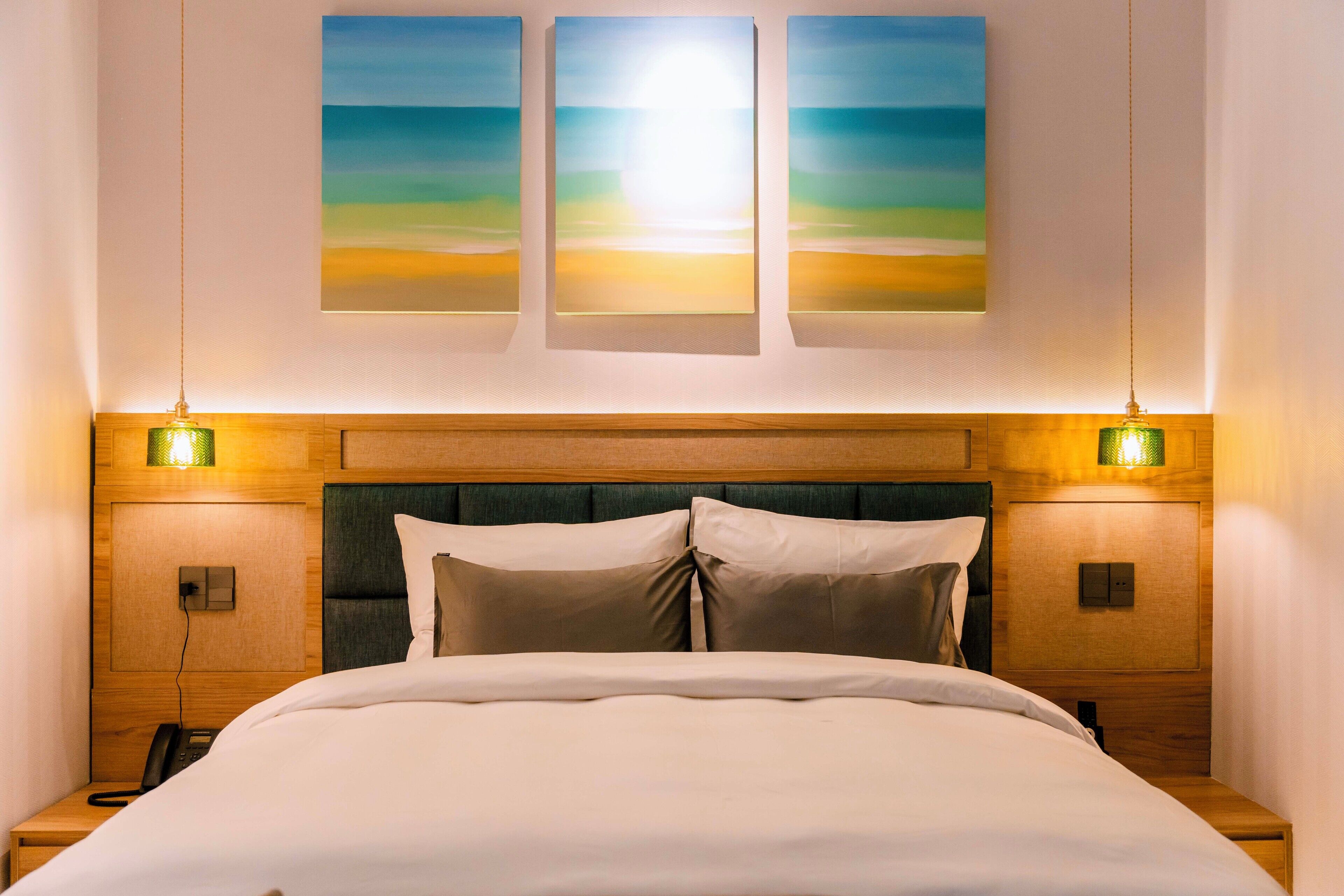 A hotel room with a large bed, two bedside tables, a telephone, and wall-mounted artwork depicting a sunset.