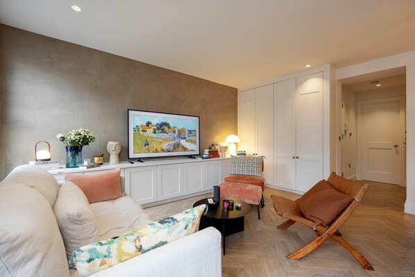 Un salon moderne avec un canapé, un fauteuil et une table basse. Un téléviseur est fixé au mur et une lampe se trouve sur la table d’appoint.