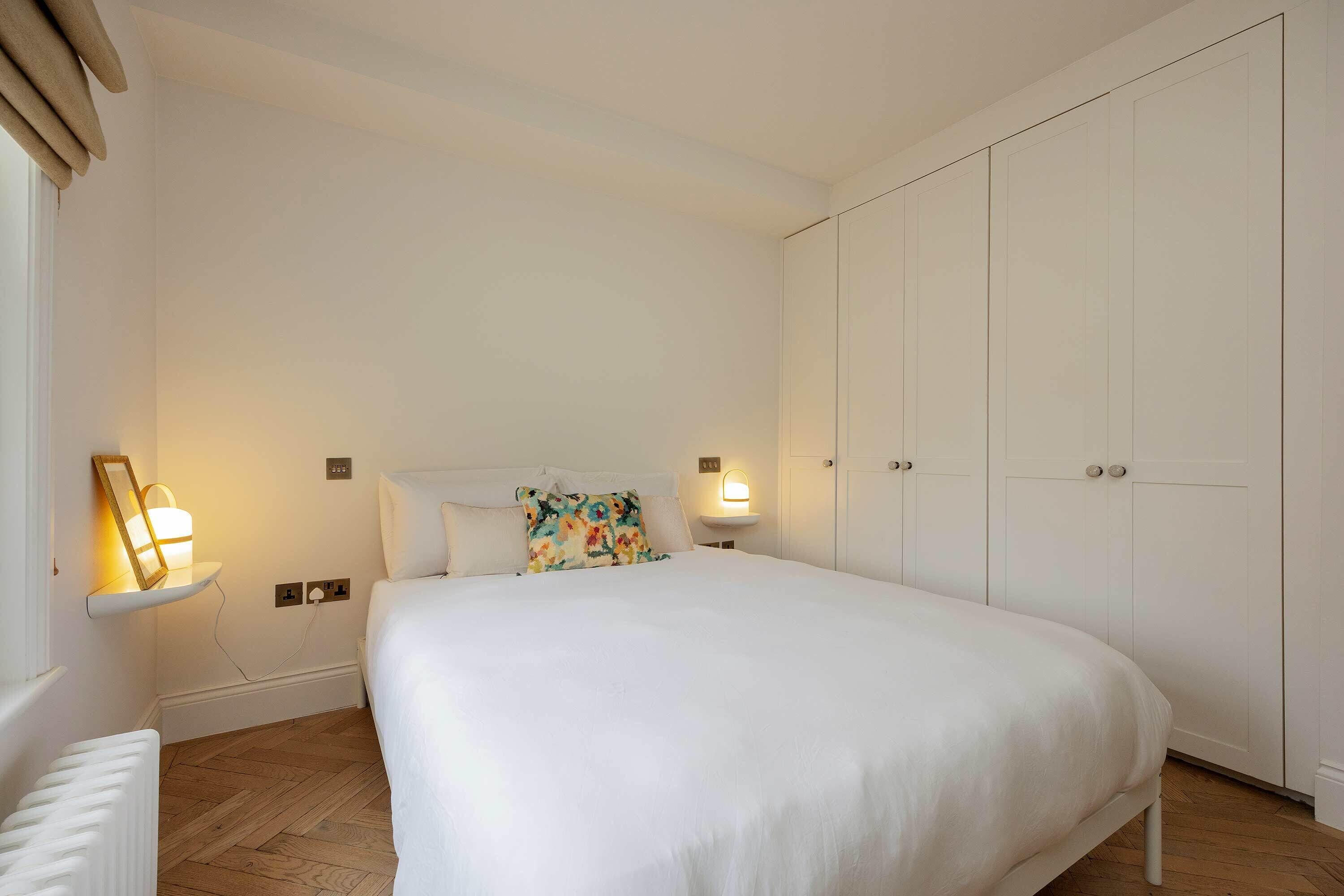 A bedroom with a large bed, white walls, wooden flooring, a wardrobe, bedside lamps, and a small shelf with a lamp.