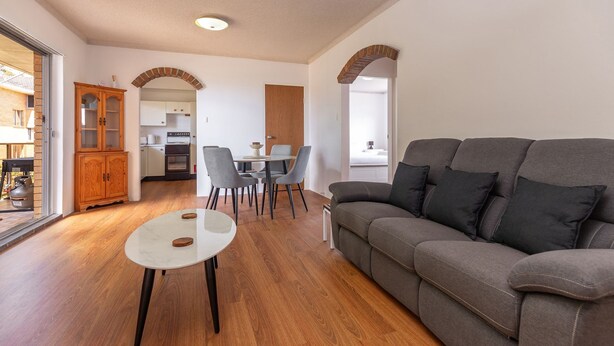 Una sala de estar moderna con un sofá gris, una pequeña mesa blanca y una zona de comedor con sillas, y una cocina al fondo.