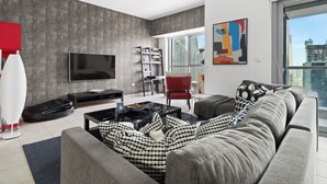 A modern living room with a grey sofa, a flat-screen TV, and a large window offering a city view. - 2304 (Dubai)