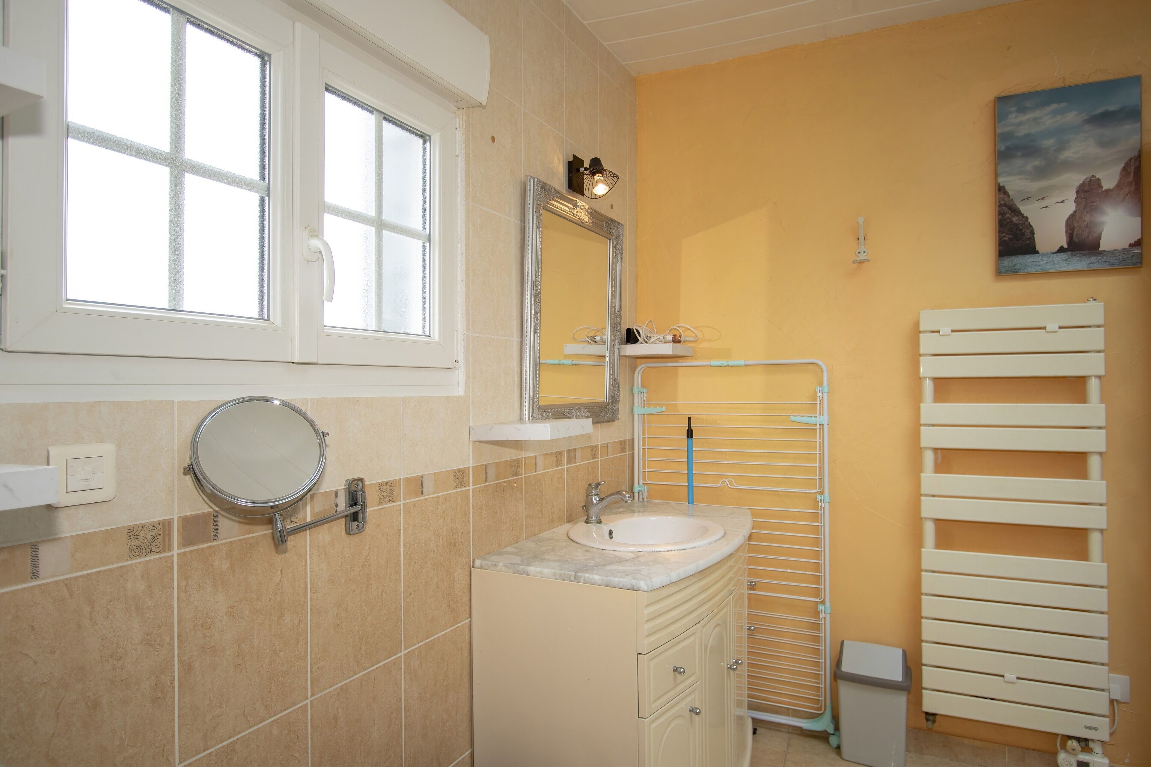Une salle de bain avec une fenêtre, un lavabo, un miroir, un porte-serviettes et une poubelle.