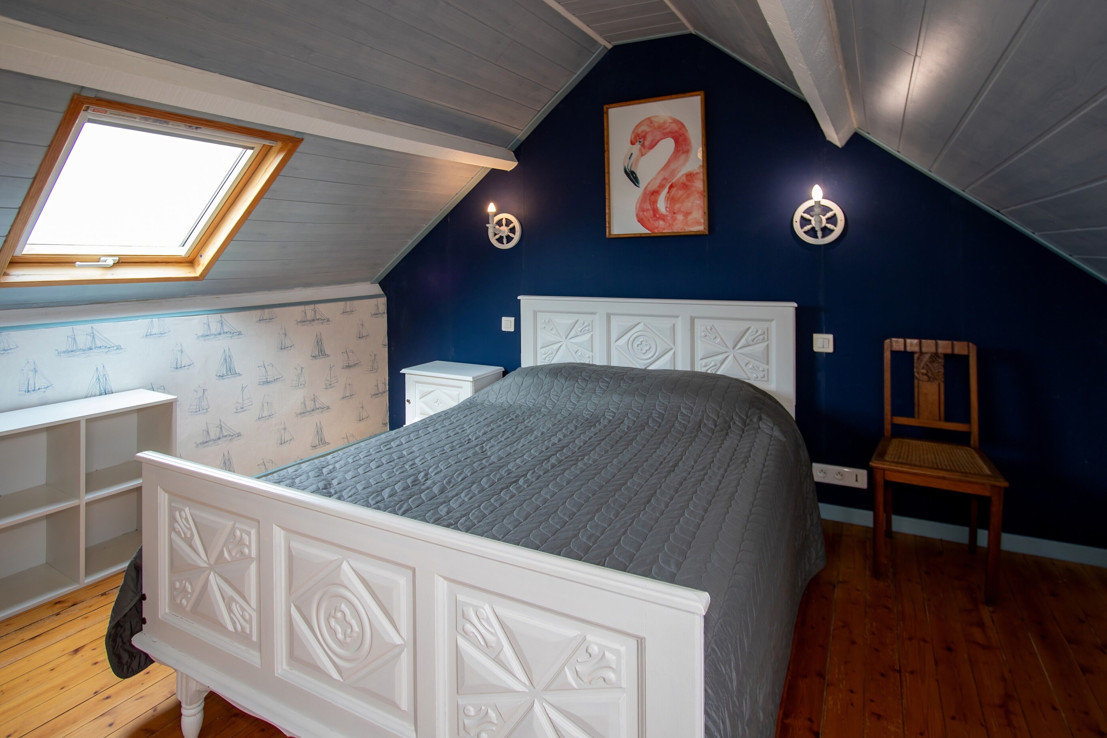 A bedroom with a sloped ceiling, a skylight, a bed with a white headboard, a chair, and a wall with a flamingo painting.