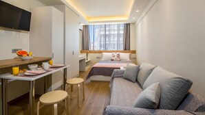Et kompakt hotelværelse med seng, sofa, skrivebord med morgenmadsprodukter og tv.