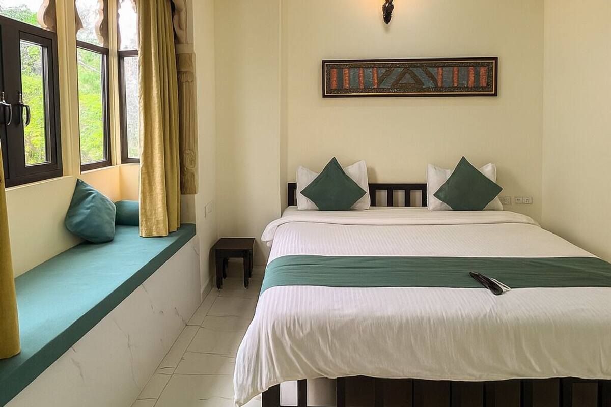 A bedroom with a bed, a window seat, a small table, and a wall-mounted light fixture.