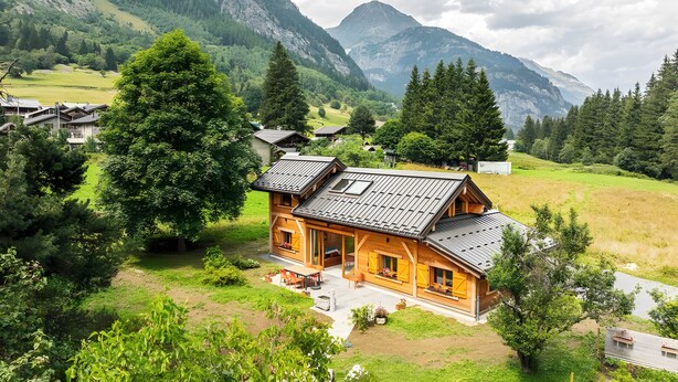 Una cabaña de madera con techo inclinado, rodeada de exuberante vegetación y montañas al fondo.