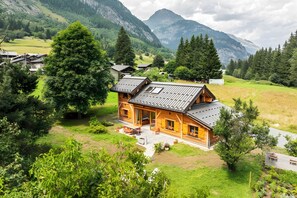 Eine Holzhütte mit geneigtem Dach, umgeben von üppiger Vegetation und Bergen im Hintergrund.