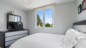 Quarto moderno com uma cama grande, uma TV em uma cômoda preta, uma janela com vista para áreas verdes e um lustre no teto.