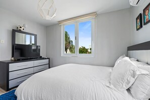 A modern bedroom with a large bed, a TV on a black dresser, a window with a view of greenery, and a ceiling light fixture. - Casa Palma Serena - Garden Retreat • Tropical Stay (Santa Isabel)