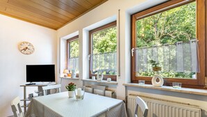 Ein Essbereich mit Tisch, Stühlen, einer Uhr, einem Fernseher und Fenstern mit Blick auf Bäume.