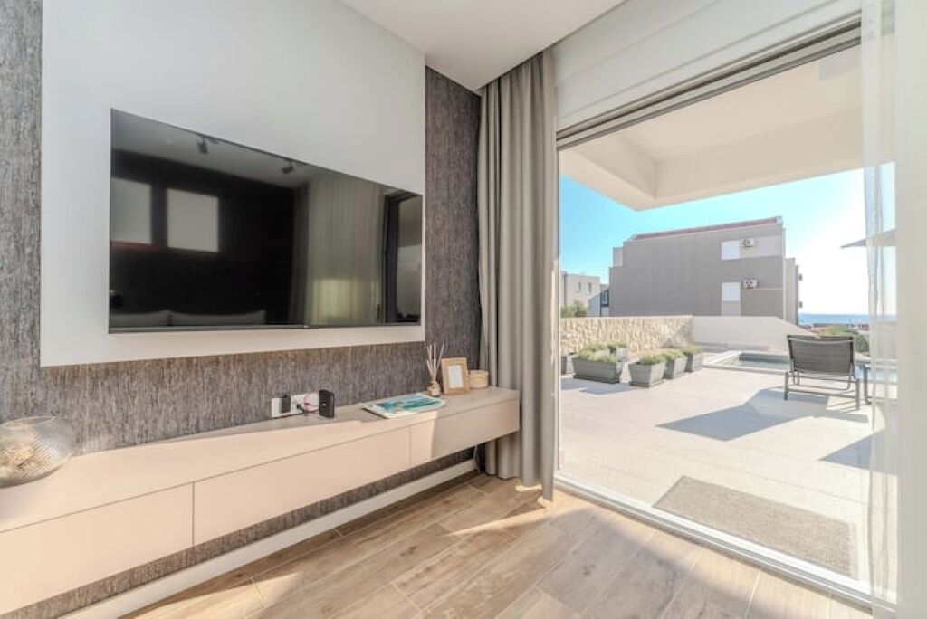 Una sala de estar moderna con un televisor de pantalla plana grande montado en la pared, piso de madera y un ventanal corredizo que da a un patio exterior con vistas a edificios y un cielo despejado.