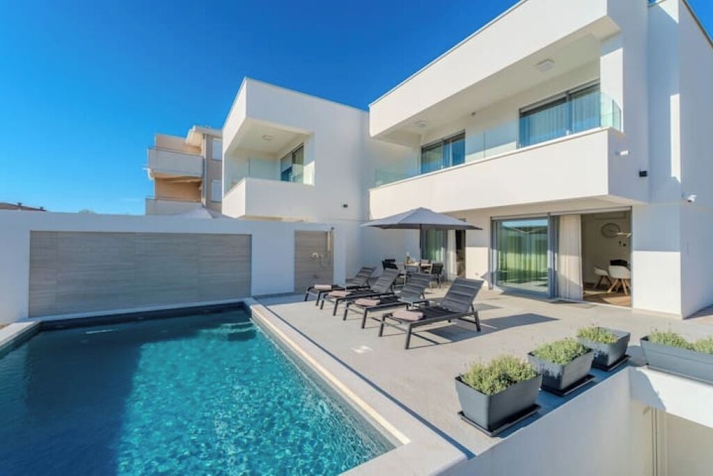 Ein modernes, zweistöckiges Haus mit Swimmingpool, Sitzgelegenheiten im Freien und einem Balkon.