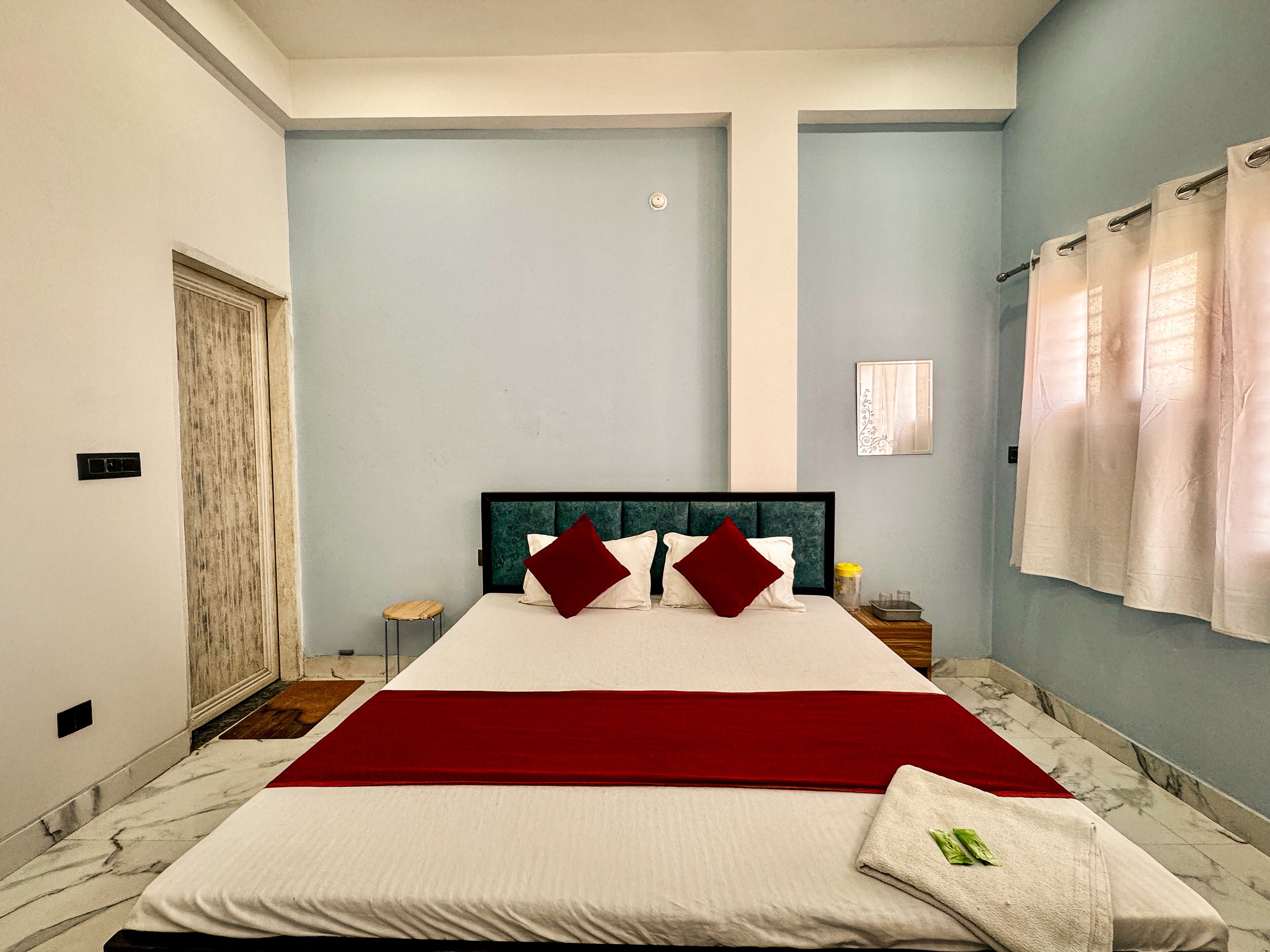 A bedroom with a large bed, a wooden stool, a nightstand, and a window with curtains.