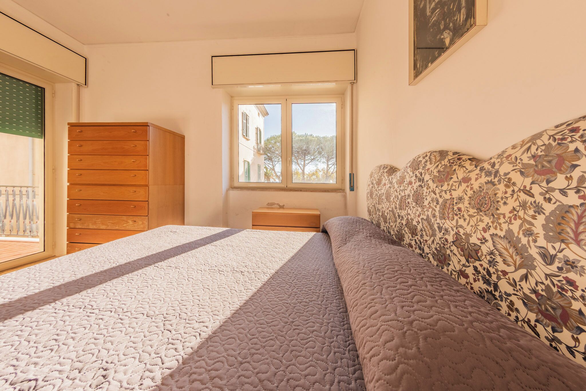 A bedroom with a bed, a dresser, a nightstand, a window, and a painting on the wall.