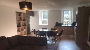 Un salon moderne comprenant un canapé, une table basse en verre, un coin repas avec une table et des chaises, une cuisine équipée de placards et une grande fenêtre laissant entrer la lumière naturelle.