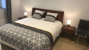 Un lit bien fait, avec une couvre-lit et des oreillers aux motifs géométriques, deux lampes de chevet, un fauteuil sombre et une fenêtre avec des rideaux.